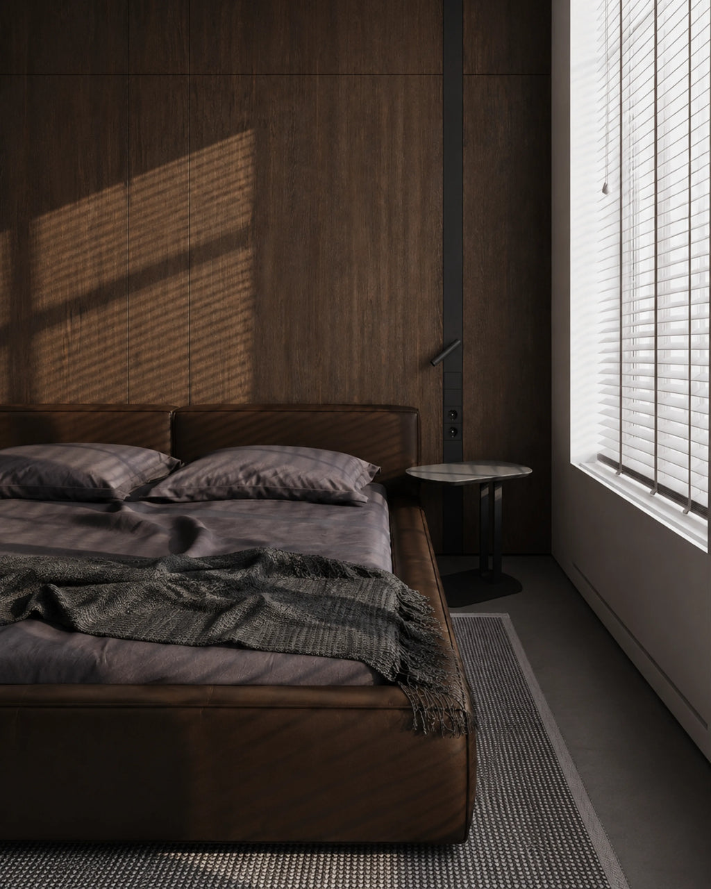 A modern bedroom scene featuring a low-profile platform bed with dark brown leather upholstery, matching dark gray bedding, and a textured throw blanket. The bed is set against a dark wood-paneled wall with warm sunlight casting dramatic shadows, next to a small side table and a window with white blinds. Saanjh bedsheets and duvet covers make your room look great. 