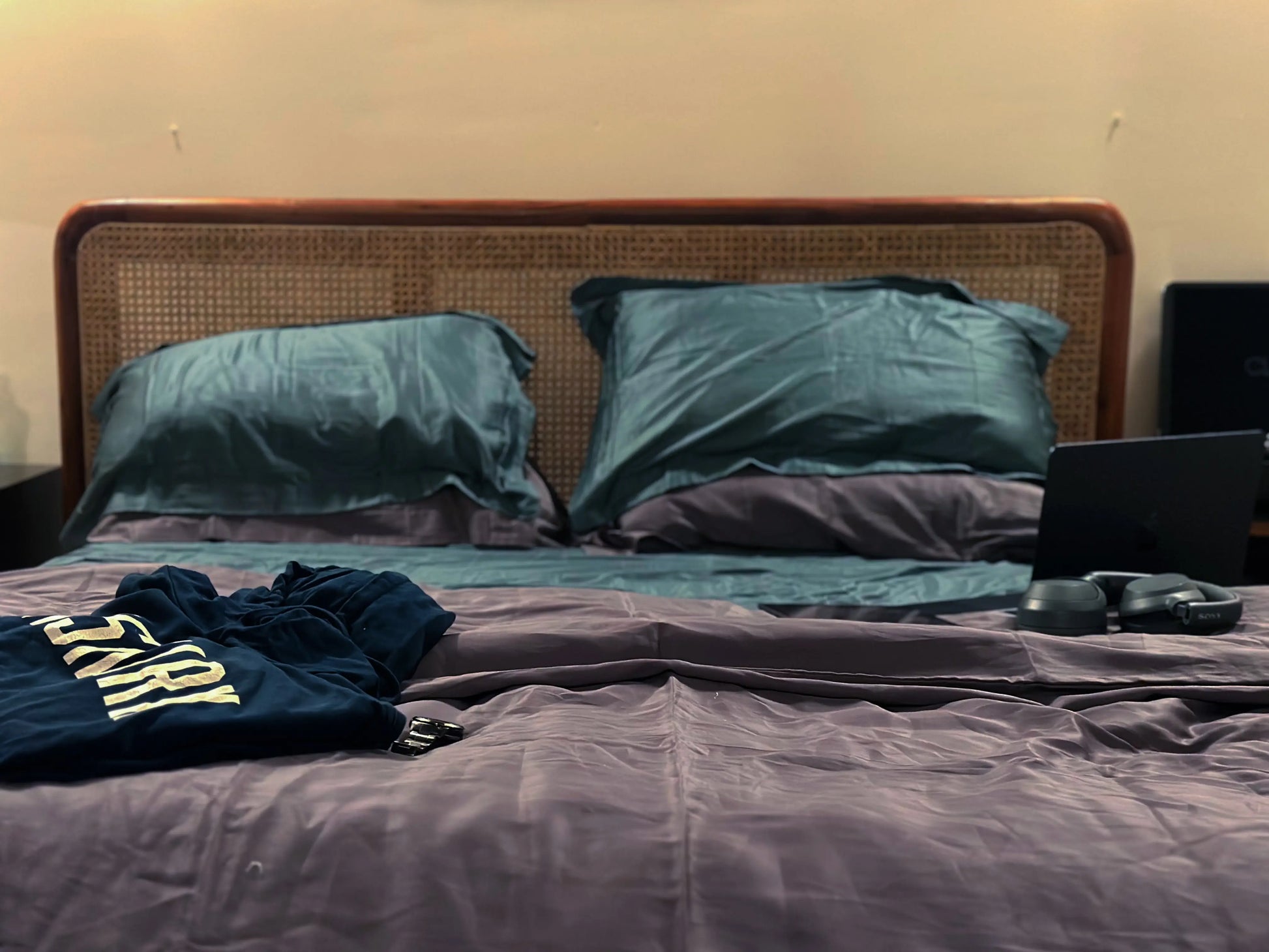 Bed with green pillows and a dark shirt on a purple blanket, against a plain wall.
