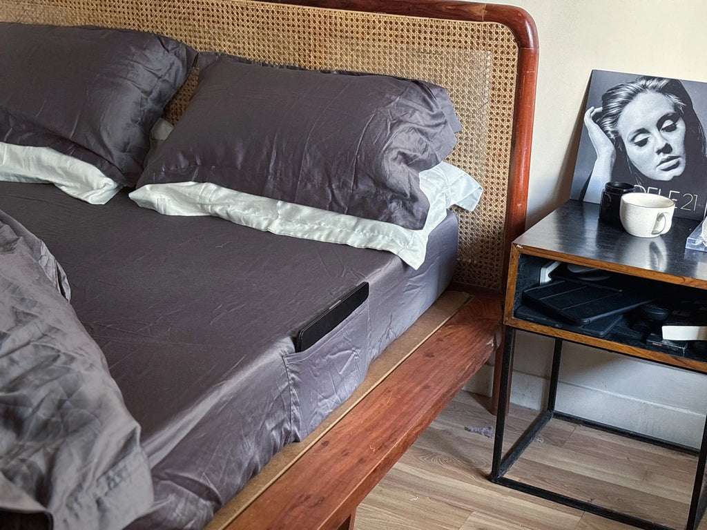 Bed with Ash gray bedding and pillows next to a nightstand with a cup and photo frame. Saanjh Homes Premium 