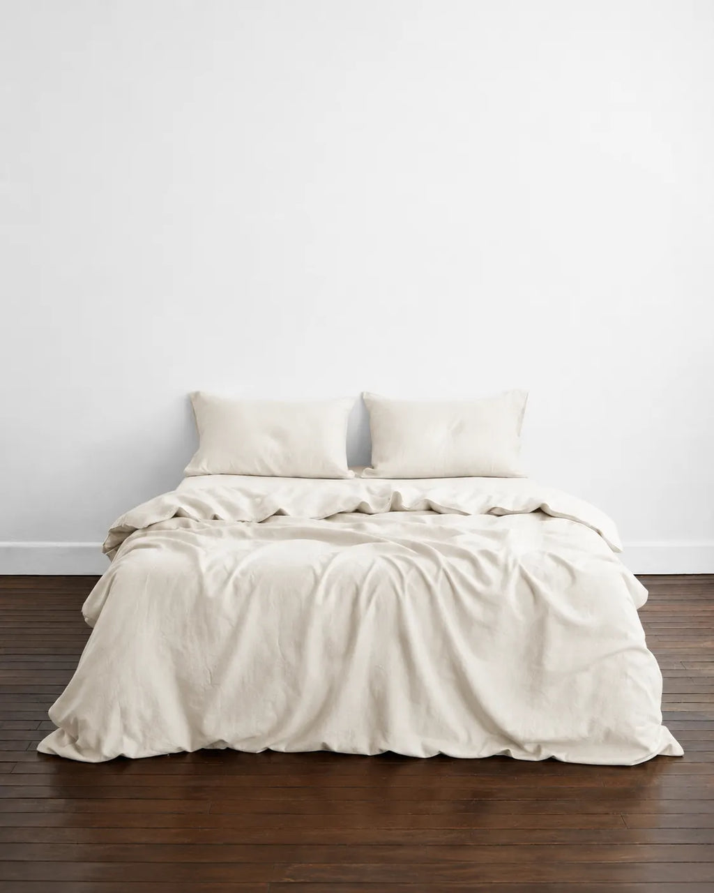 A neatly made bed with rumpled saanjh creme bedding and two matching pillows, set against a plain white wall on a dark wood floor.