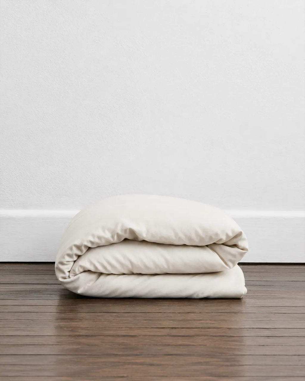 A folded saanjh homes' cream duvet or comforter sits on a dark wood floor against a white wall.