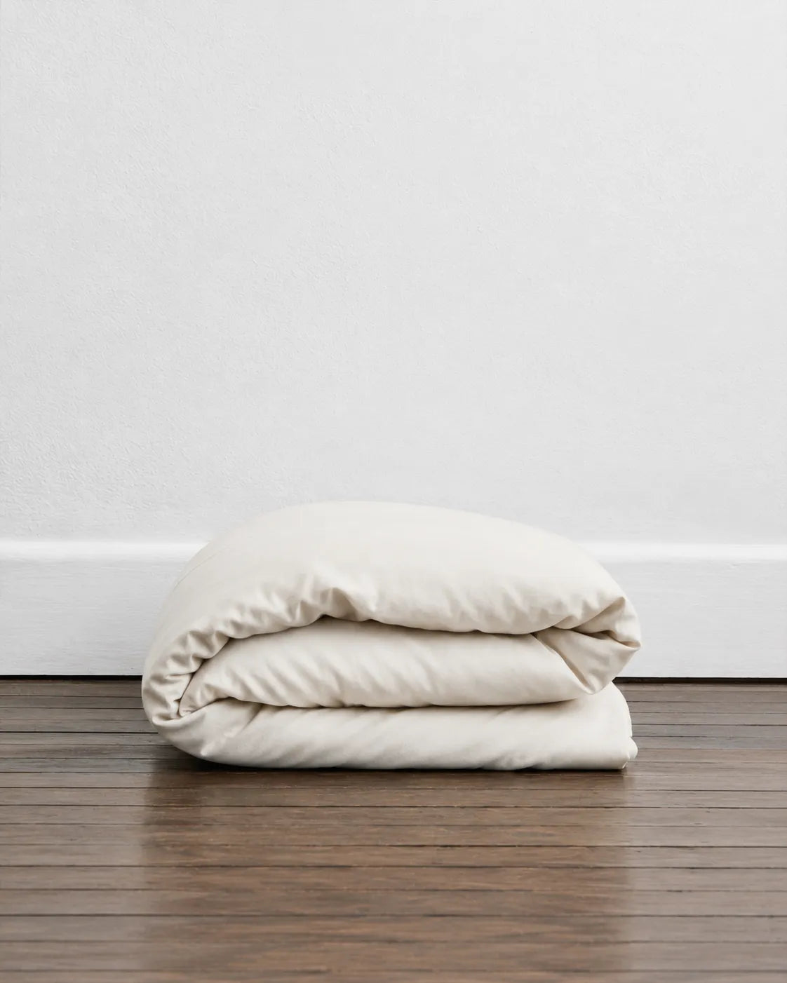A folded saanjh homes' cream duvet or comforter sits on a dark wood floor against a white wall.
