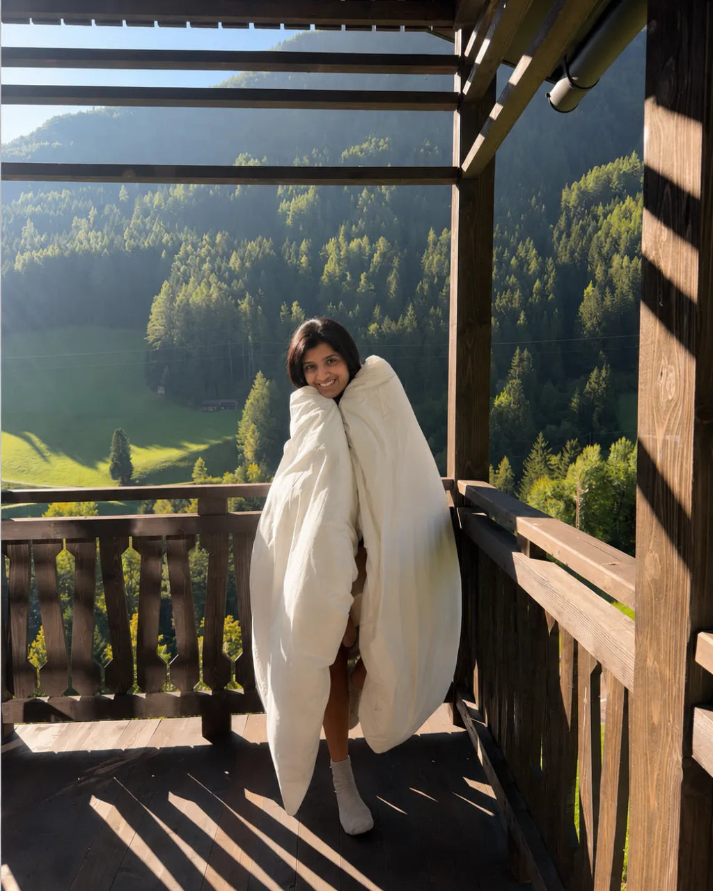 A woman wrapped in a large, plush creme duvet stands on a wooden balcony overlooking a lush green mountain valley.