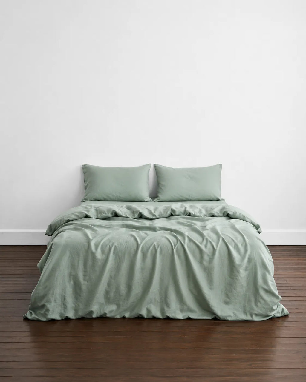 A bed dressed in a saanjh soft mint bedding set with two matching pillowcases, styled on a dark wood floor against a plain white wall.