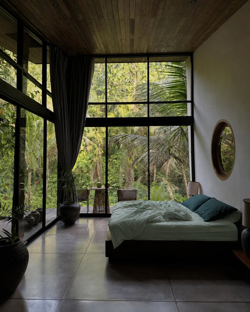 A modern bedroom with a low platform bed, mint bedding, and petrol pillows from saanjh homes, set in a room with a wood ceiling, dark gray tiled floor, and a large floor-to-ceiling window looking out onto a lush green forest.