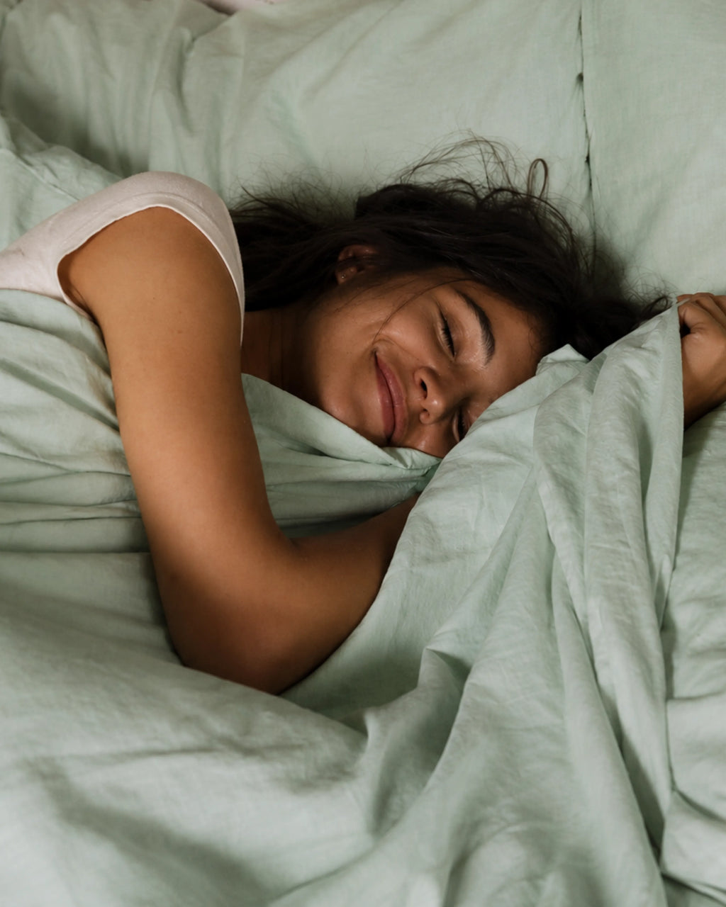 A woman sleeping soundly in bed, wrapped in saanjh homes' mint bedding. 
