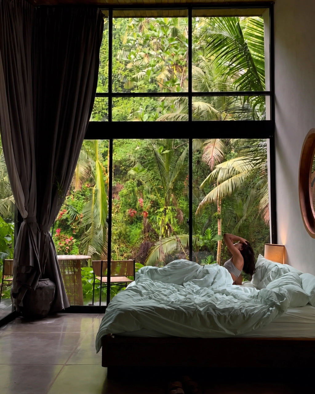 A person relaxes in bed with rumpled saanjh homes' mint bedding, positioned under a large black-framed window that overlooks a lush tropical green jungle view. Dark gray curtains are tied back on the left, and a warm bedside lamp glows on the right.