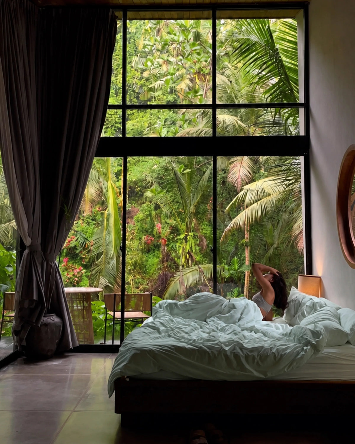 A person relaxes in bed with rumpled saanjh homes' mint bedding, positioned under a large black-framed window that overlooks a lush tropical green jungle view. Dark gray curtains are tied back on the left, and a warm bedside lamp glows on the right.