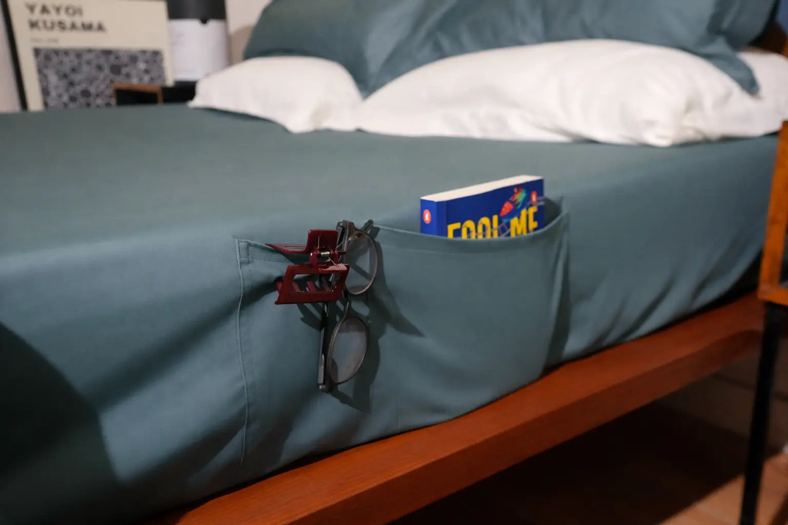 Bed with a pocket holding sunglasses and a book titled 'Fool Me Twice' against a blue bedspread.