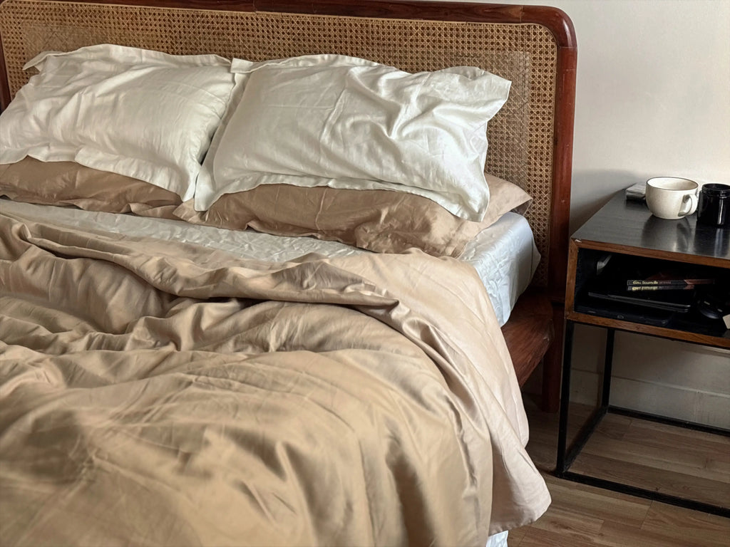 Bed with beige bedding and white pillows next to a nightstand with a cup and remote control.