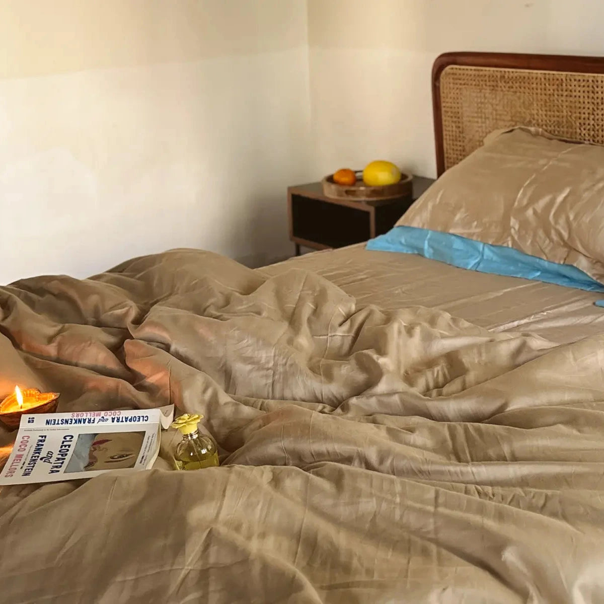 Bed with beige bedding and a wooden headboard, nightstand with fruit, and lit candle. From Saanjh Homes Aamras collection